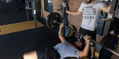 A strong man lifts weights on a bench while a trainer assists in a modern gym setup.