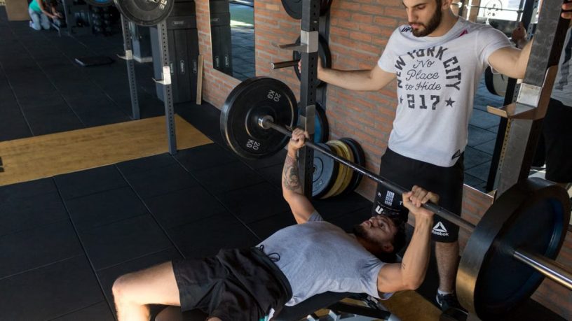 A strong man lifts weights on a bench while a trainer assists in a modern gym setup.
